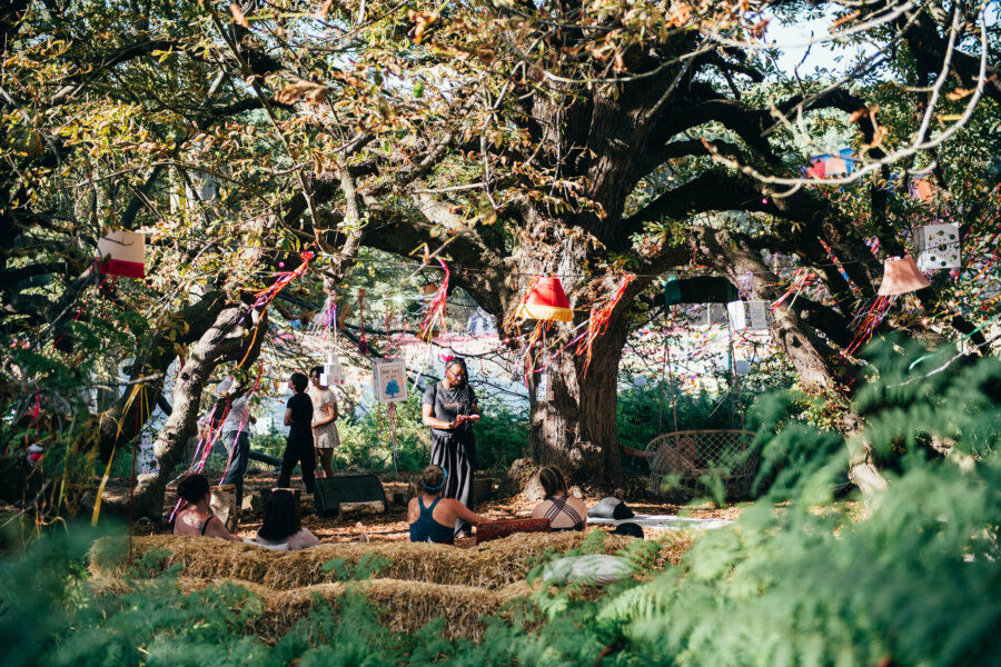 people sitting under a large tree called 'the tree of life' at latitude festival. the tree is decorated with streamers and lampshades. 