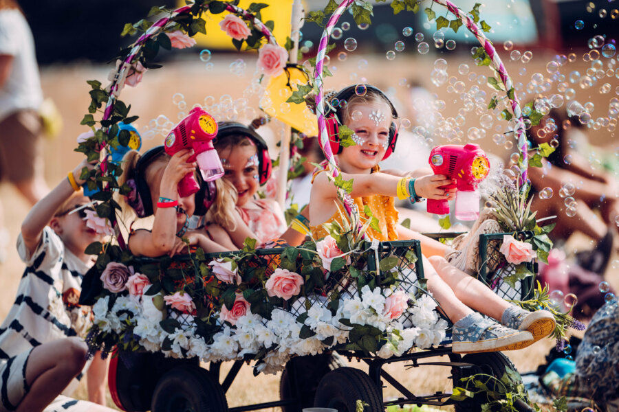 three young children having sun in a trolley, which is covered in flowers. the children are blowing bubbles with a bubble gun.