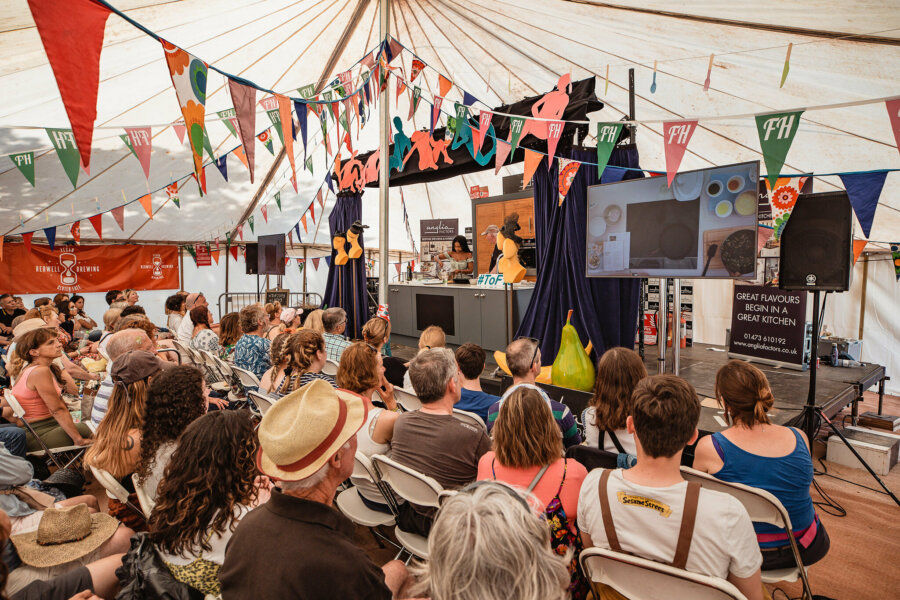 a full Theatre of Food tent at Latitude. Rows of people are sat facing a stage where a cooking demonstration is taking place.